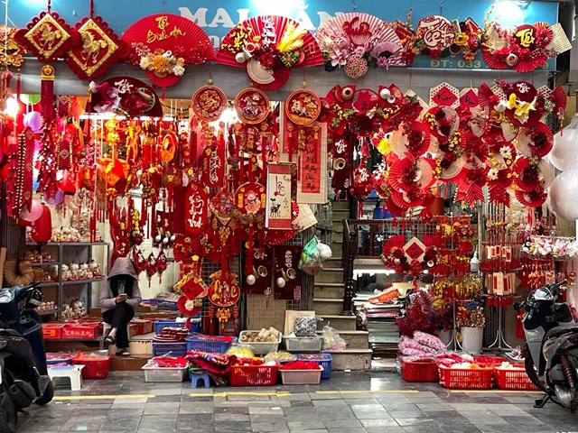 Lọt thỏm giữa sắc đỏ rực rỡ của h&agrave;ng ng&agrave;n m&oacute;n đồ trang tr&iacute;, người b&aacute;n lặng lẽ lướt điện thoại trong cảnh vắng kh&aacute;ch hiếm thấy.